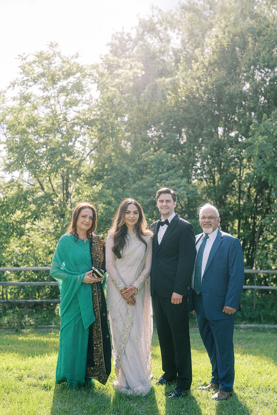A formal family portrait during a garden wedding ceremony, featuring a bride in a traditional embroidered saree, a groom in a classic tuxedo, and elegantly dressed family members, set against a lush, sunlit backdrop.
