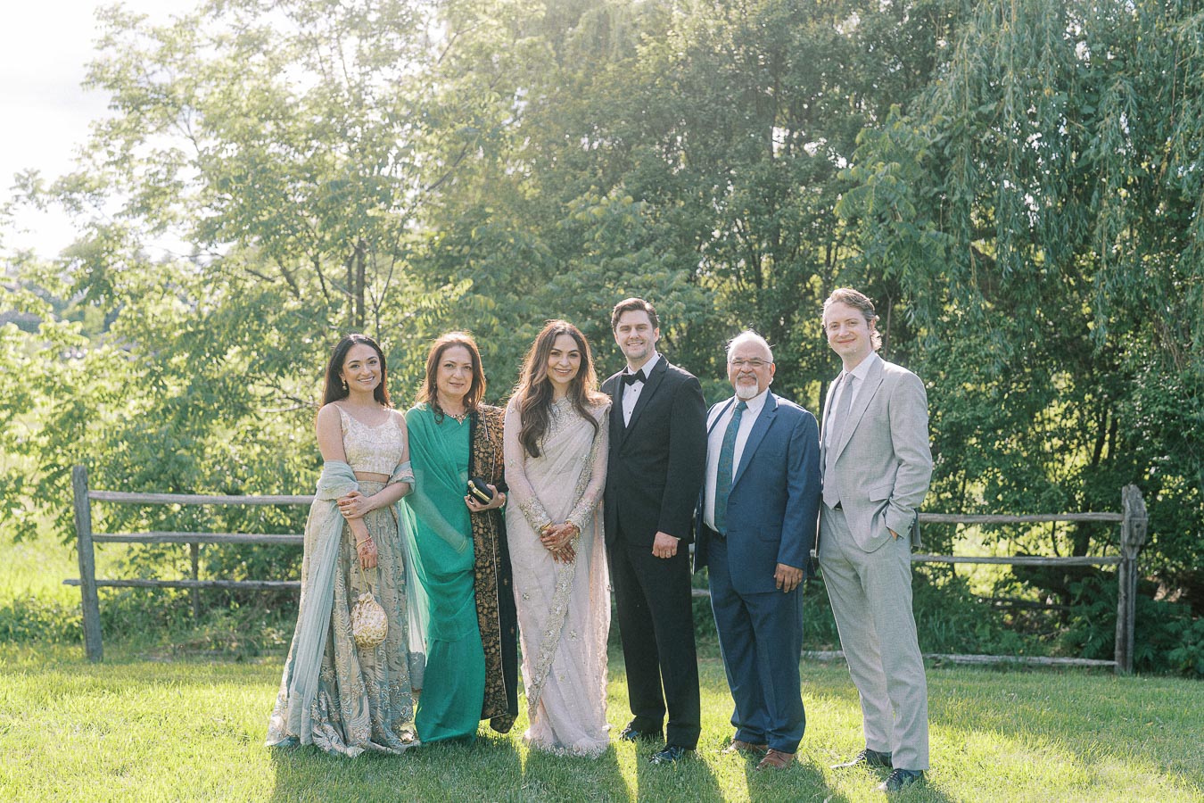 A group of six people standing together outdoors, dressed in formal attire comprising suits and elegant dresses, with lush green trees and a wooden fence in the background. They appear to be celebrating a special occasion in a garden setting on a sunny day.