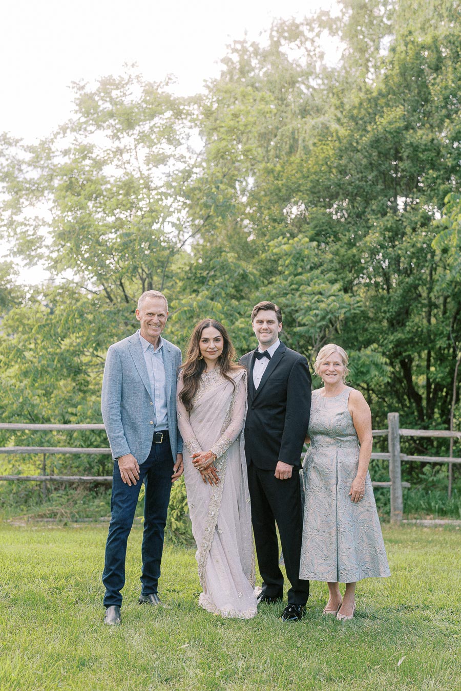 A group of four people standing outdoors on a sunny day, dressed in formal attire, including a man in a light blue blazer, a woman in an elegant saree, another man in a tuxedo, and a woman in a silver dress, with green trees and a wooden fence in the background.