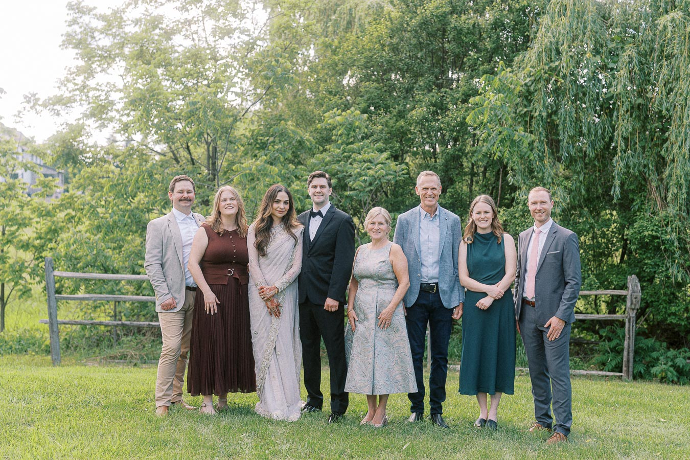 A group of eight people dressed in formal and semi-formal attire posing for a photograph outdoors in a lush green garden setting.