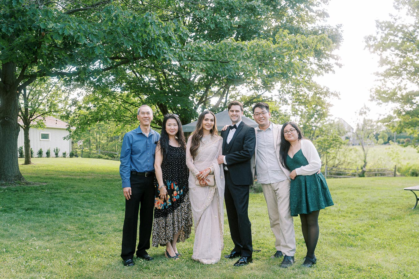 A group of six people smiling and posing in an outdoor setting with lush green trees and grass. One person is dressed in a traditional sari, while another is in a tuxedo, ideal for a wedding or formal occasion. The rest of the group is casually dressed, creating a vibrant and joyful scene of celebration and togetherness.