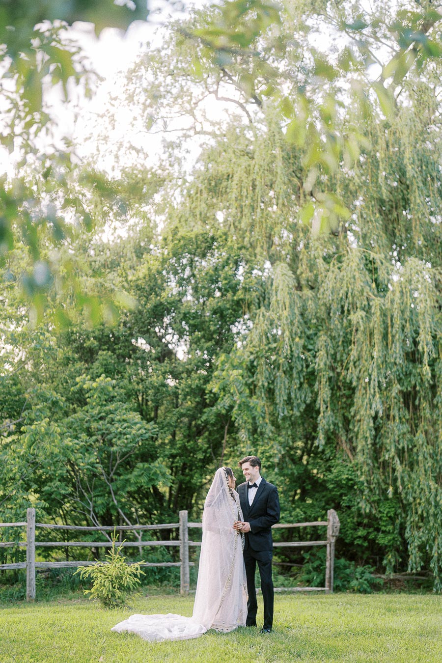 Bride and groom standing in a lush green garden, embracing in front of a wooden fence and leafy trees, with the bride in a flowing white gown and veil, and the groom in a black tuxedo.