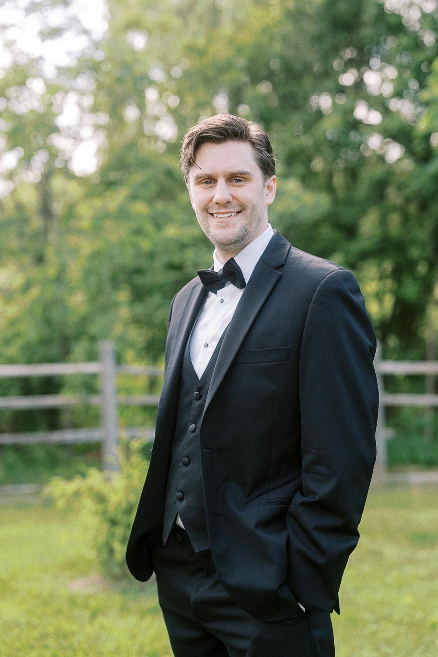 Smiling man in a tuxedo with a bow tie, standing outdoors with green trees in the background.