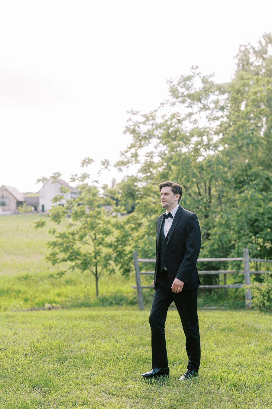 A man in a formal black tuxedo walks confidently across a lush green field, with a rustic fence and trees in the background, under a clear sky.