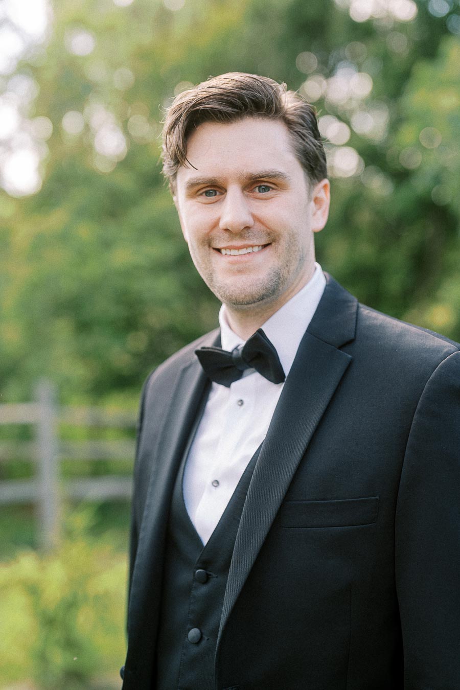 Young man in a formal black tuxedo smiling outdoors with a blurred greenery background.