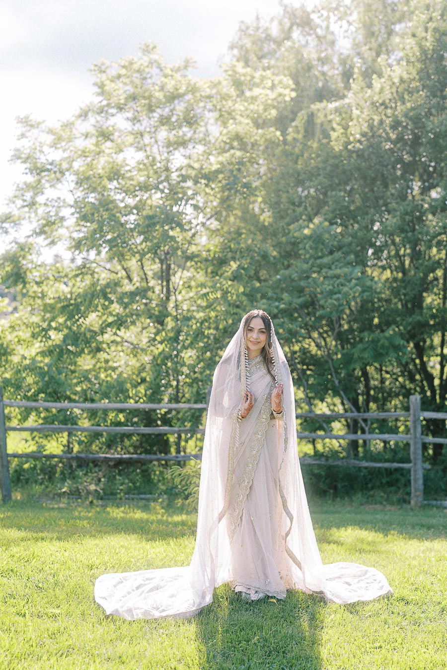 Bride in an elegant wedding outfit with a veil standing in a sunlit garden with lush green trees and wooden fence in the background.