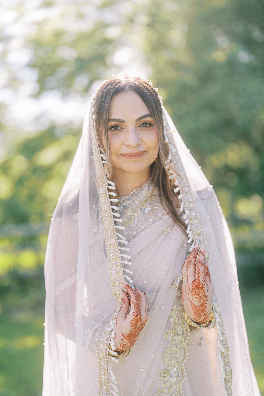 A smiling woman in traditional bridal attire with intricate embroidery and a veil, showcasing detailed henna designs on her hands, set against a natural, sunlit outdoor background.