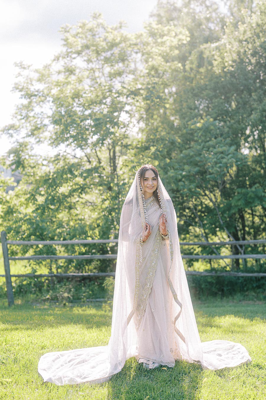 Bride in elegant white bridal saree with delicate embroidery, hands adorned with henna, standing in a sunlit garden setting with green trees.