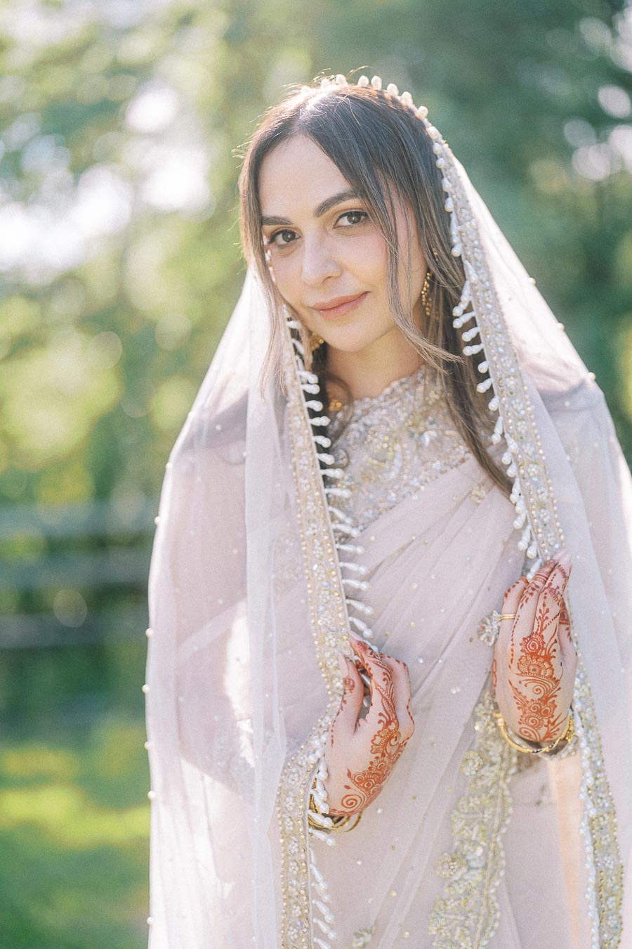 Elegant woman in traditional attire with intricate henna designs on her hands, adorned in a embellished veil, standing outdoors with a blurred green background.