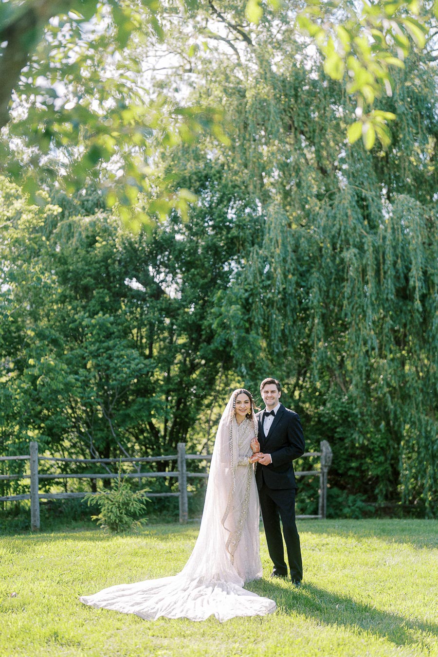 Bride and groom standing outdoors in a serene garden setting, with the bride in an elegant gown and veil, and the groom in a black tuxedo, surrounded by lush green trees and sunlight filtering through the leaves.
