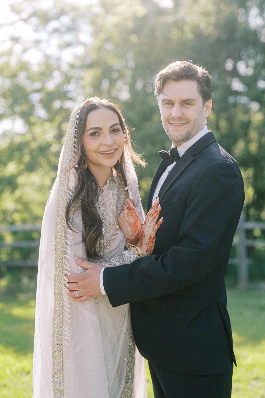 Bride and groom smiling on their wedding day, with the bride in a traditional embroidered dress and the groom wearing a tuxedo, standing outdoors with greenery in the background.