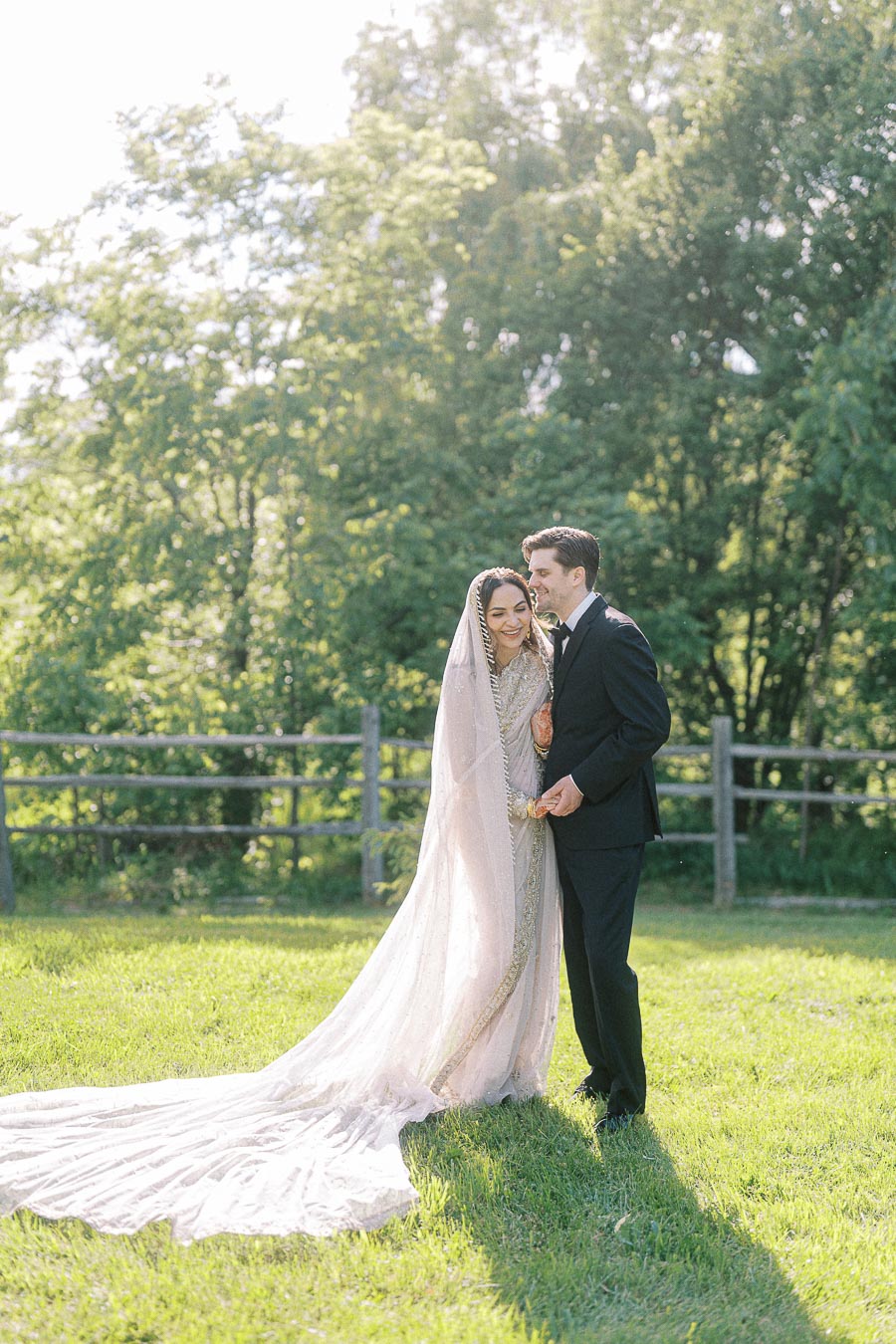 Bride and groom standing in a sunlit garden, wearing traditional wedding attire, surrounded by lush greenery and a rustic wooden fence.