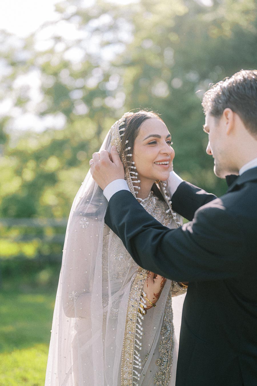 A bride in an elegant, embroidered veil smiling as her groom adjusts it during a sunny outdoor wedding ceremony.