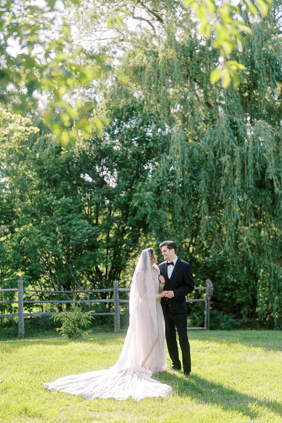 Bride and groom in elegant attire stand together in a sunlit garden with lush greenery, capturing a romantic outdoor wedding scene.
