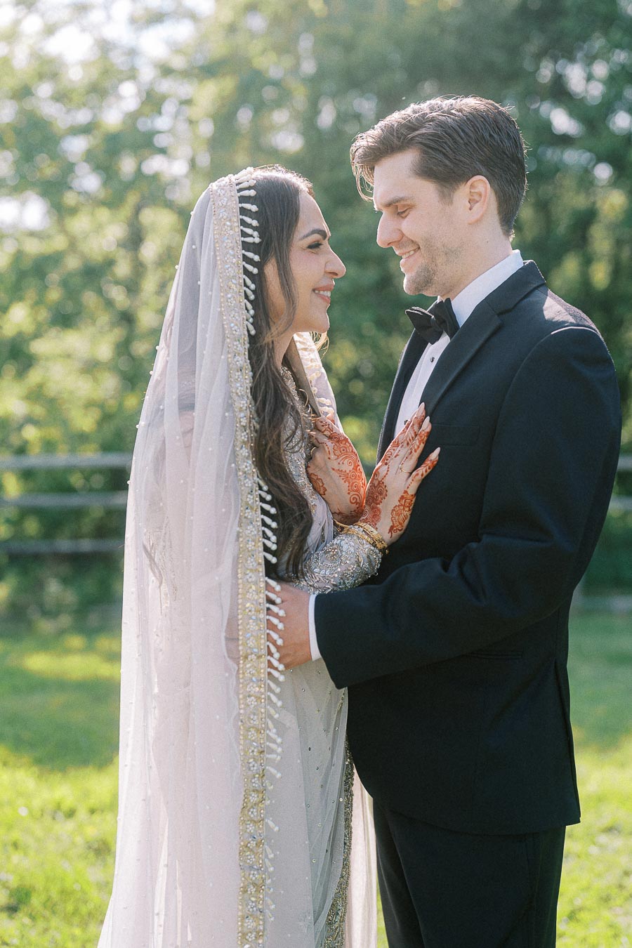 A bride in a beautiful embellished white bridal outfit adorned with henna on her hands, standing closely with the groom in a classic black tuxedo in a lush green garden, both gazing into each other's eyes with joy.