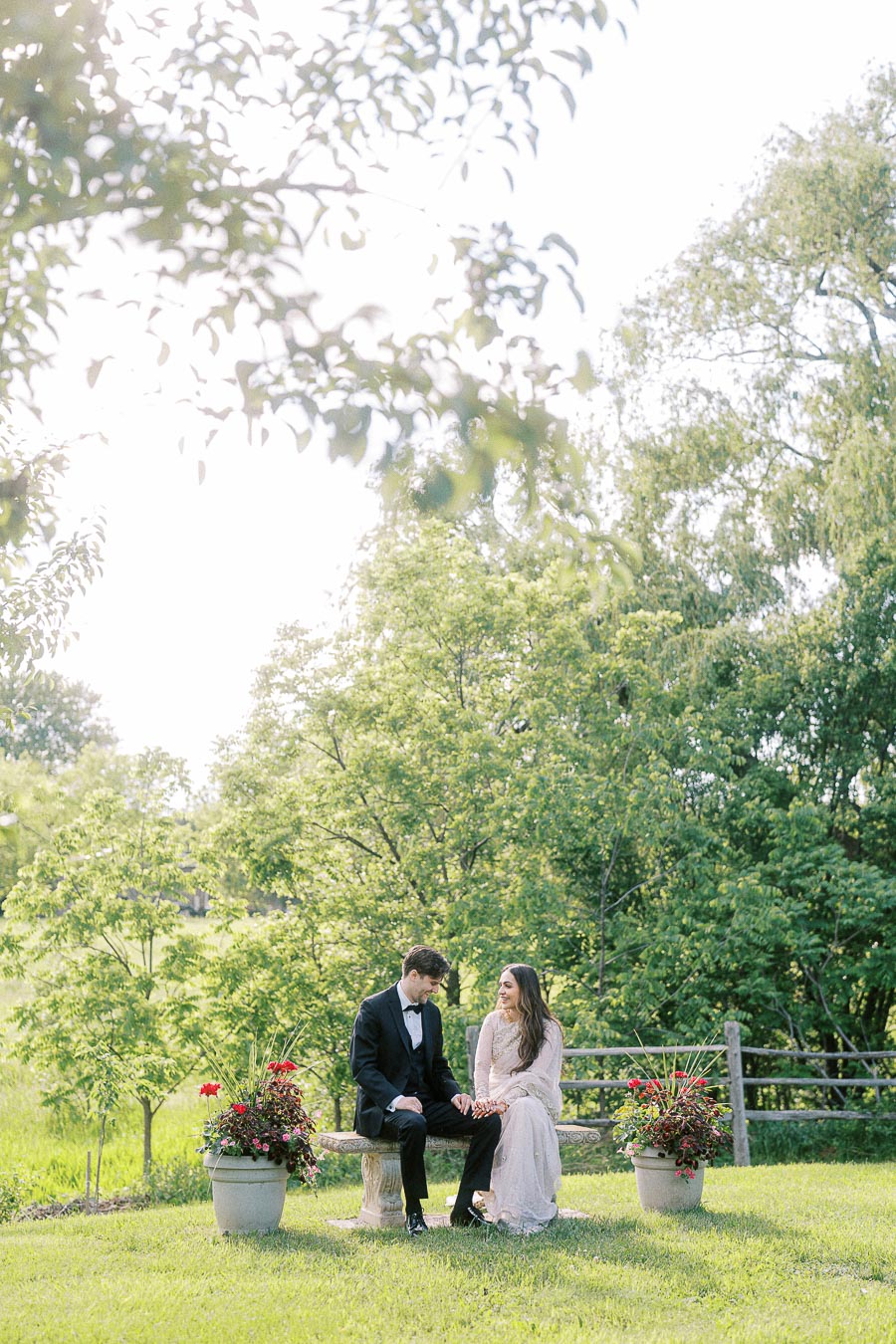 A couple dressed in formal attire sitting on a stone bench in a lush green garden with trees and blooming flowers.