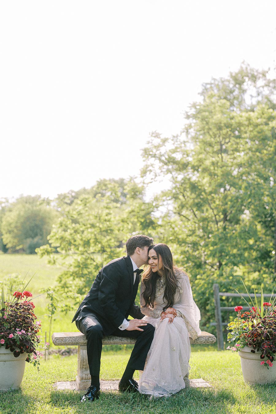 A couple in wedding attire sits on a garden bench, surrounded by lush greenery and floral arrangements, sharing an intimate moment with the groom whispering to the bride.