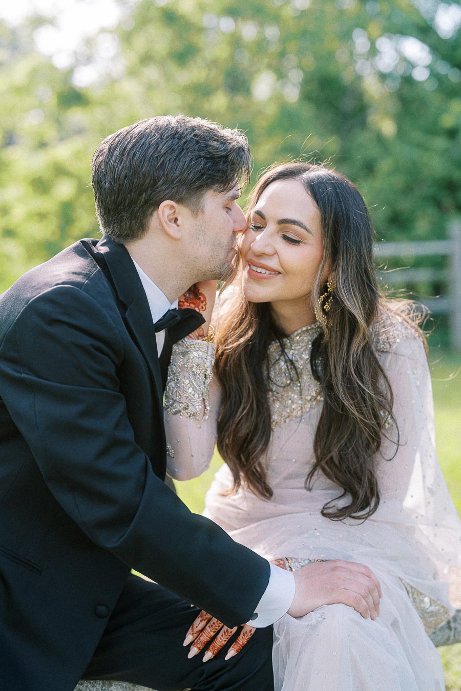 A couple enjoying a romantic moment outdoors, with the man in a black tuxedo gently kissing the woman's cheek as she smiles, wearing an ornate white dress and showing intricate henna designs on her hand.