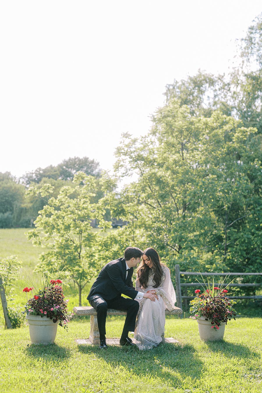 A couple sitting on a bench in a sunny garden setting, surrounded by lush greenery and potted flowers, sharing an intimate moment.
