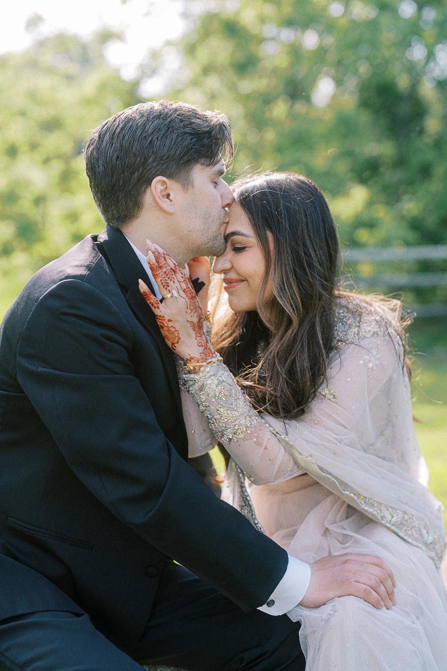 A couple in formal attire sharing an intimate moment outdoors, with the man in a black suit gently kissing the woman's forehead. The woman is wearing an elegant dress adorned with intricate details and displaying henna designs on her hands. The background features lush greenery.