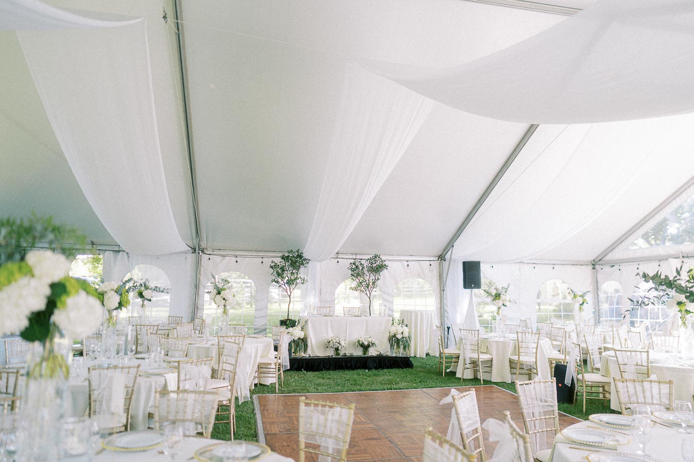 Elegant wedding reception setup under a white marquee tent with draped ceilings, decorated with round tables adorned with white tablecloths and golden chairs, floral centerpieces, and a small stage for the head table, creating a sophisticated outdoor celebration ambiance.