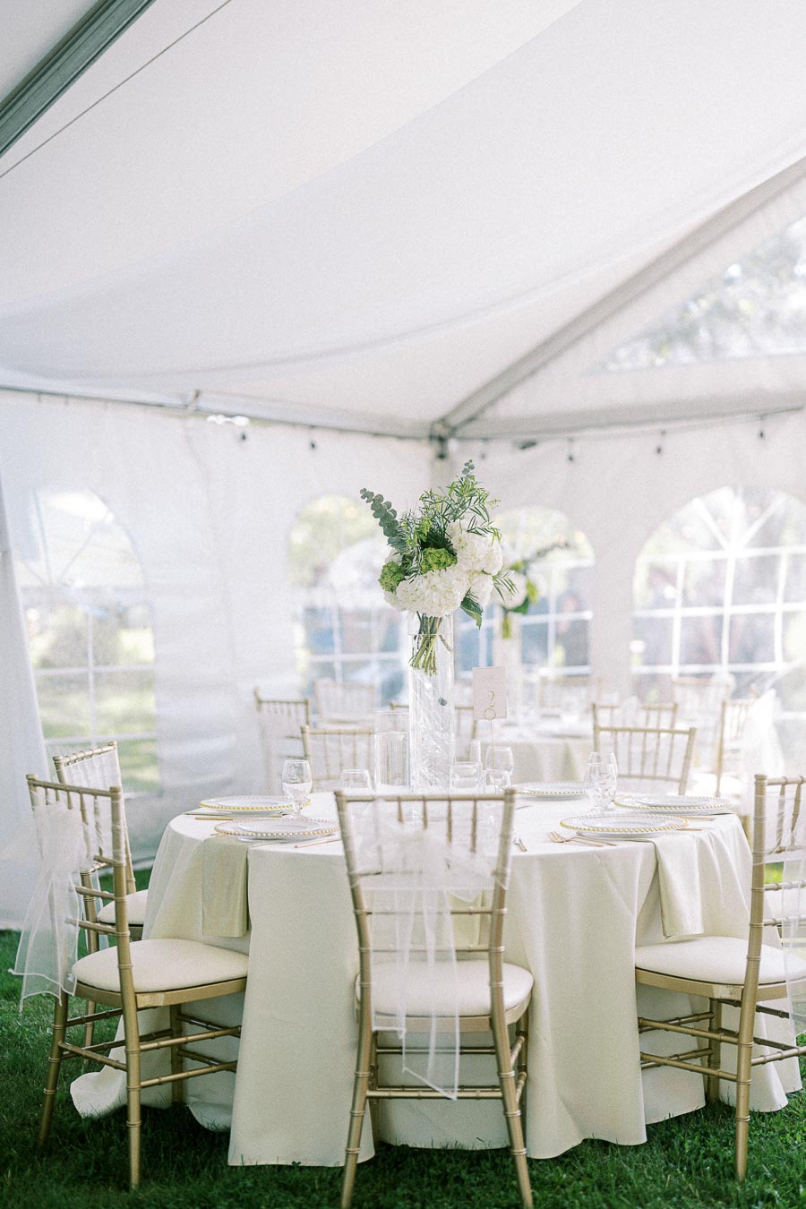 Elegant wedding reception setup under a white tent with round tables, gold chiavari chairs, white tablecloths, and tall floral centerpiece arrangements.