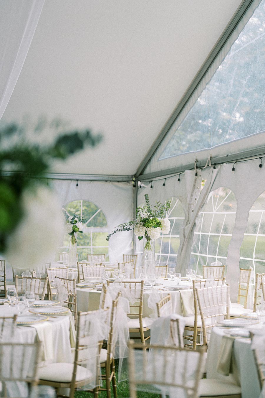 Elegant wedding reception tent with white table settings and gold chairs featuring floral centerpieces.