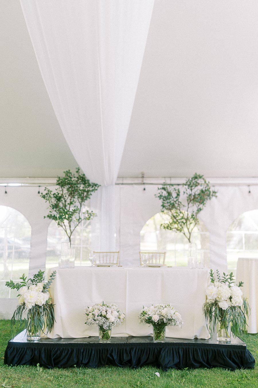 Elegant outdoor wedding reception table under a white tent, adorned with lush greenery, floral arrangements, and a classic white tablecloth, creating a sophisticated and romantic setting.