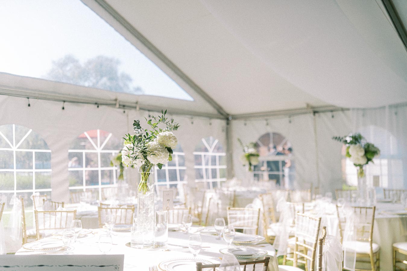 Elegant wedding reception in a bright, sunlit tent with round tables adorned with white flowers and gold chairs.