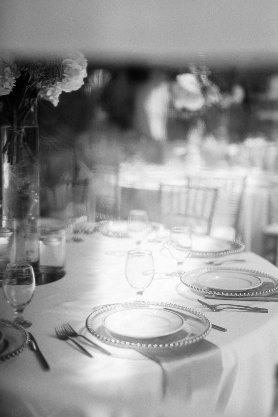 Elegant black and white image of a sophisticated table setting with crystal plates, wine glasses, and floral centerpiece on a white tablecloth in a blurred soft focus, ideal for weddings or special events.
