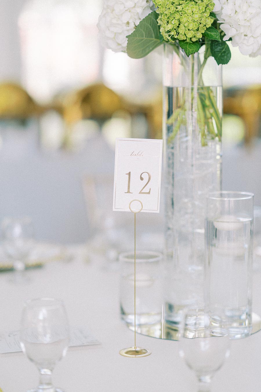 Elegant wedding table setting with table number 12 in gold, featuring a tall glass vase filled with green and white hydrangeas, surrounded by candles and glassware on a white tablecloth.
