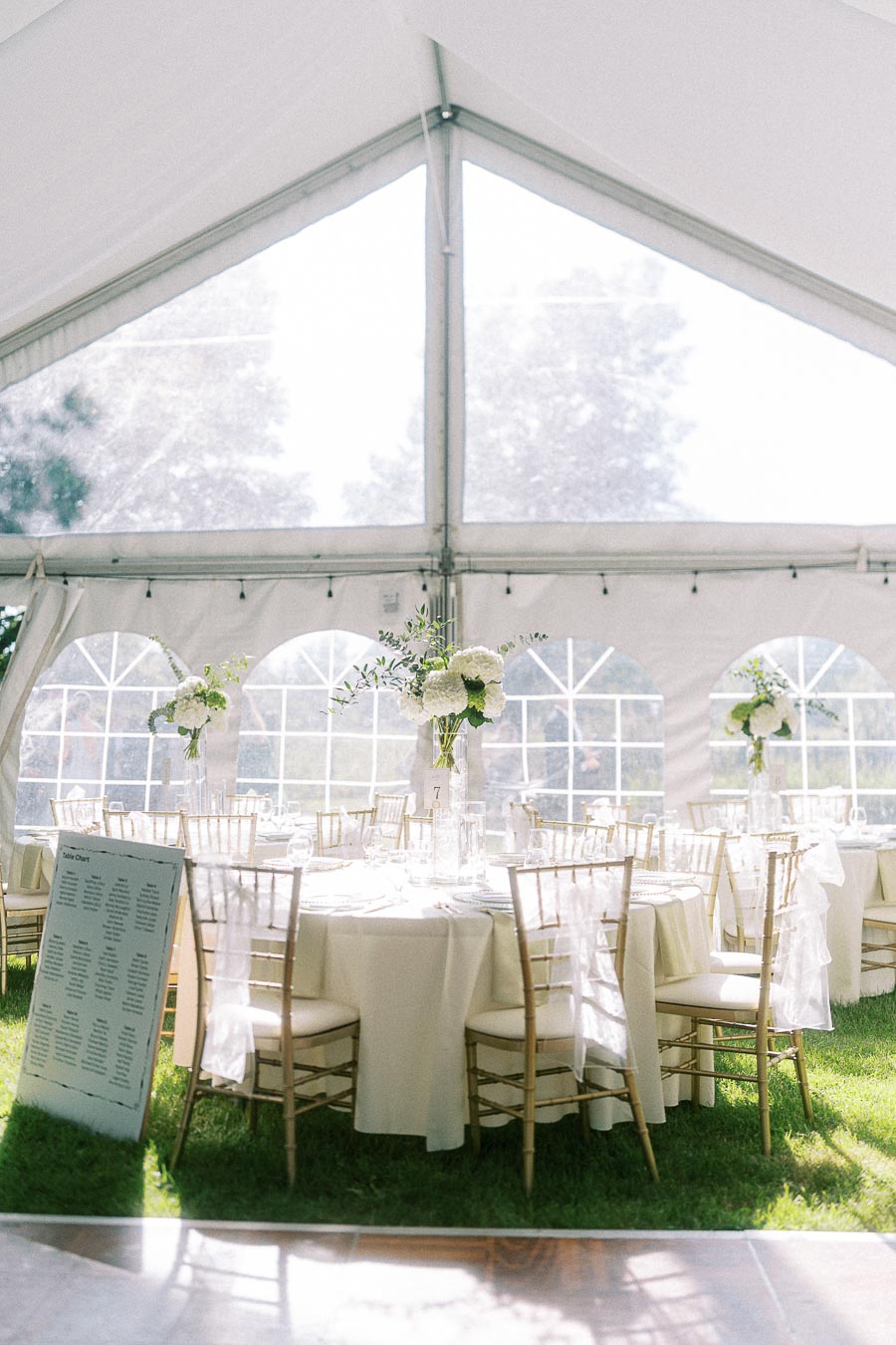 Elegant wedding reception setup inside a white tent, featuring round tables with white tablecloths, gold chiavari chairs, and floral centerpieces, creating a bright and sophisticated ambiance.