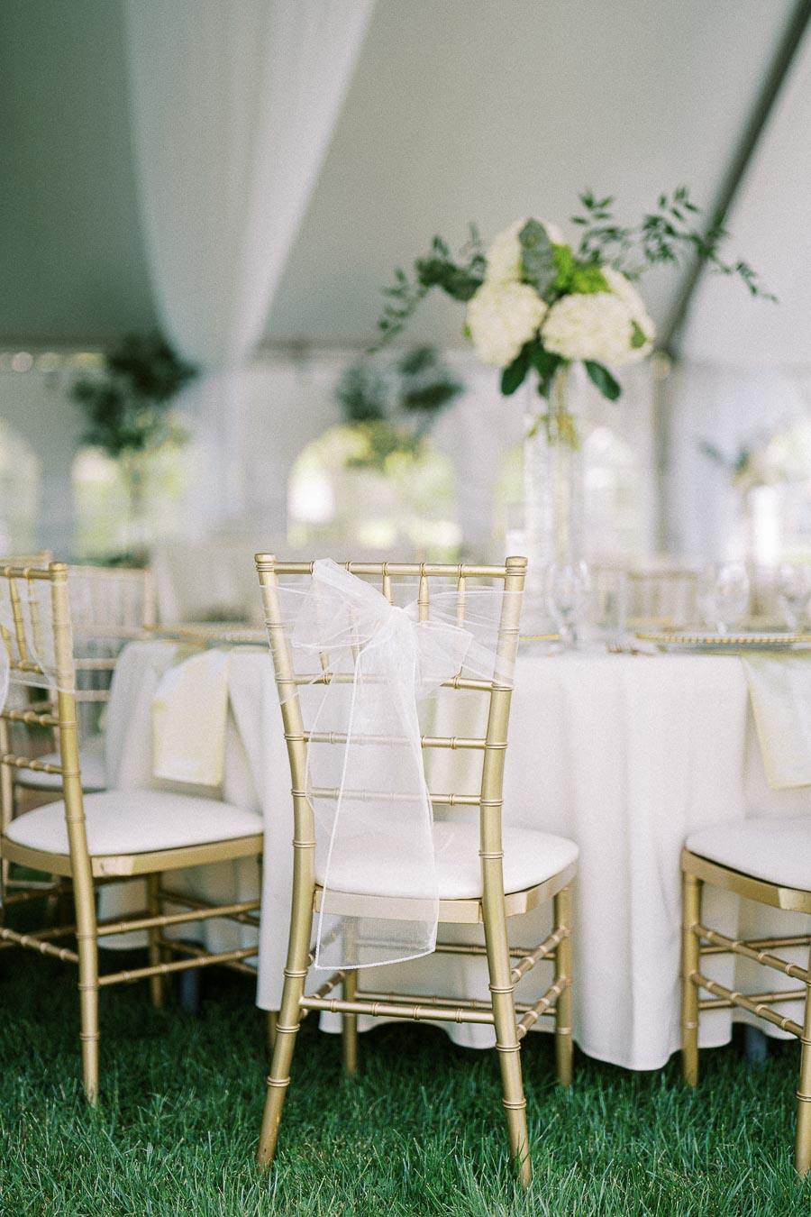 Elegant outdoor wedding reception setup under a tent, featuring white-draped round tables, gold Chiavari chairs with sheer white bows, and a tall centerpiece with white flowers and greenery.
