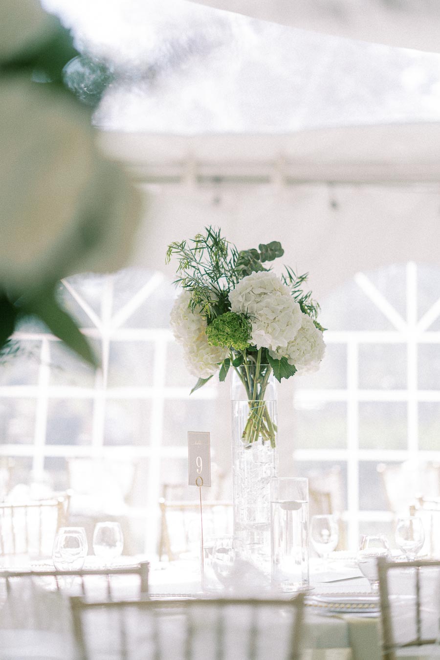 Elegant wedding reception table setting with a tall glass vase centerpiece featuring white and green hydrangeas, surrounded by clear glassware and a number nine table card, under a softly lit marquee tent.