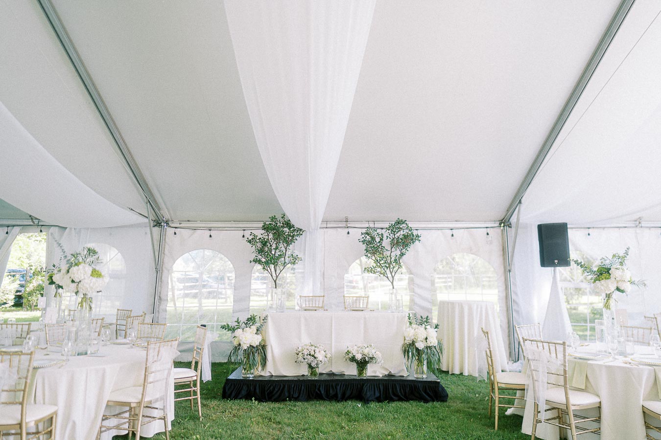 Elegant wedding reception tent with white drapery, gold chairs, floral centerpieces, and greenery accents.