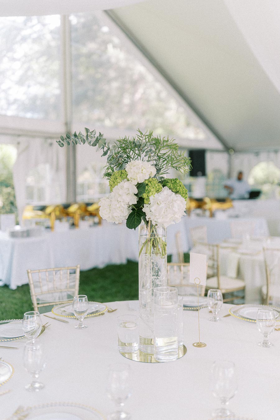 Elegant wedding reception table setup with white hydrangea and greenery centerpiece in a clear vase, surrounded by fine glassware and gold-accented tableware under a white event tent.