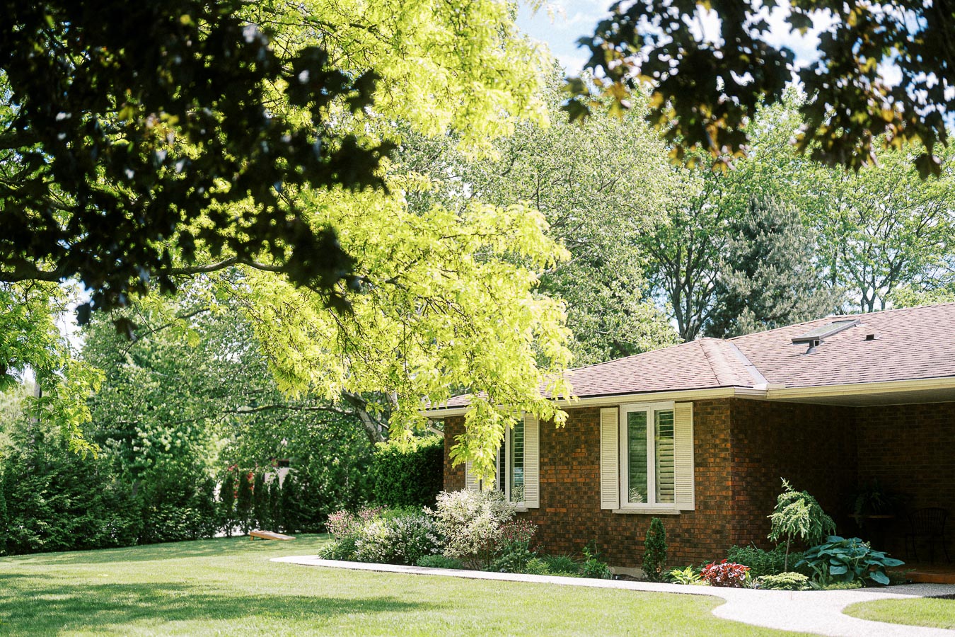 Alt text: Front view of a brick house surrounded by lush green trees and a well-manicured lawn, showcasing a serene garden setting under a sunny sky.