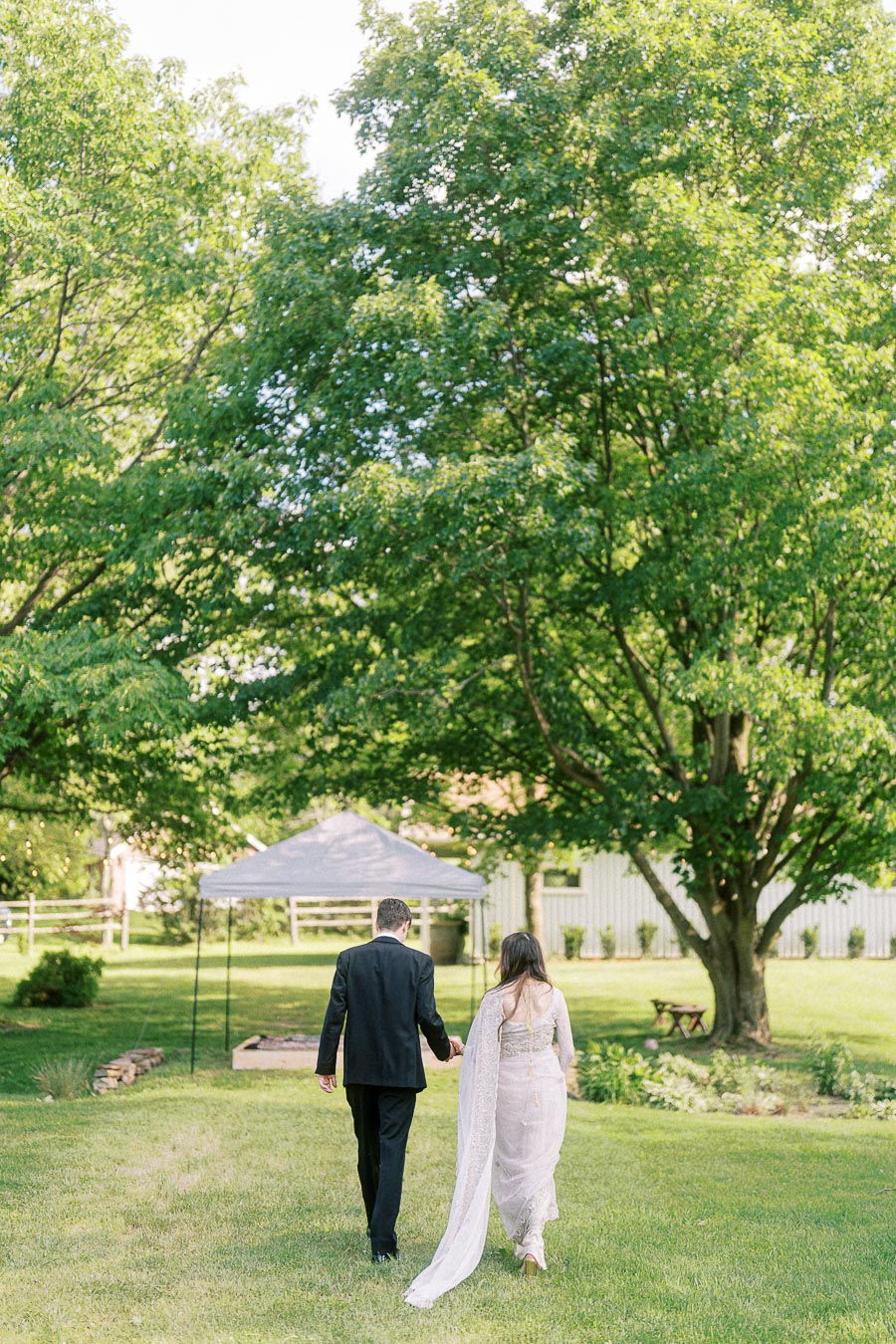 A couple walking hand in hand in a picturesque garden setting with lush green trees and a white event tent in the background.