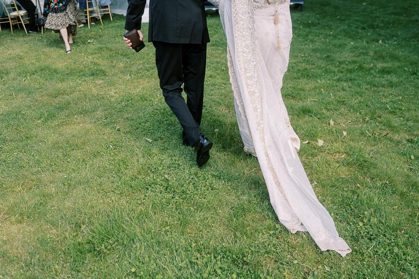 A couple walking hand in hand on green grass, with the woman wearing an elegant embellished wedding dress and the man in a formal black suit, captured at an outdoor event.