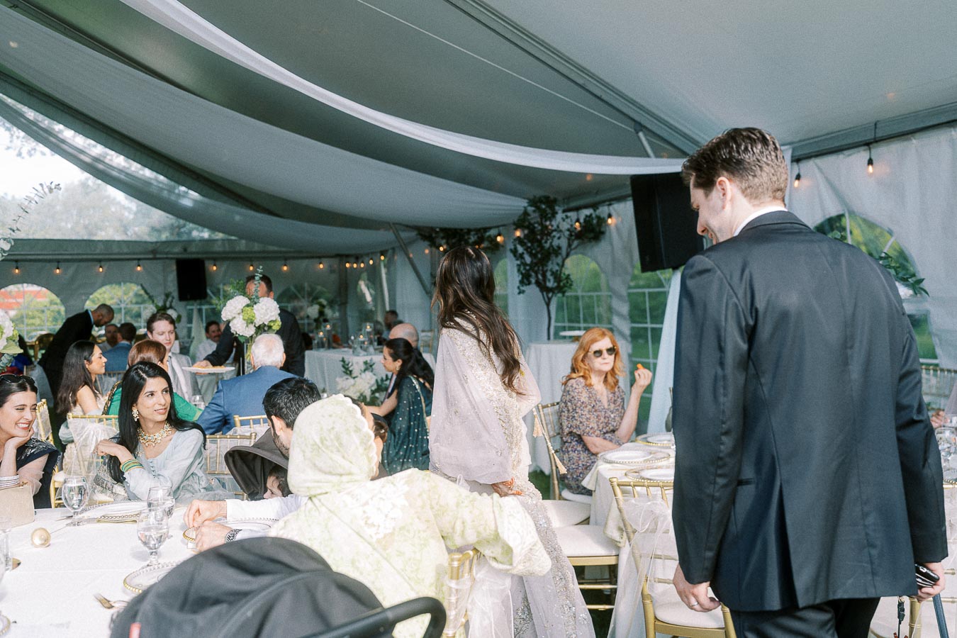 A vibrant wedding reception under a tent with elegantly dressed guests sitting at decorated tables engaged in conversation, featuring floral centerpieces and warm ambient lighting.