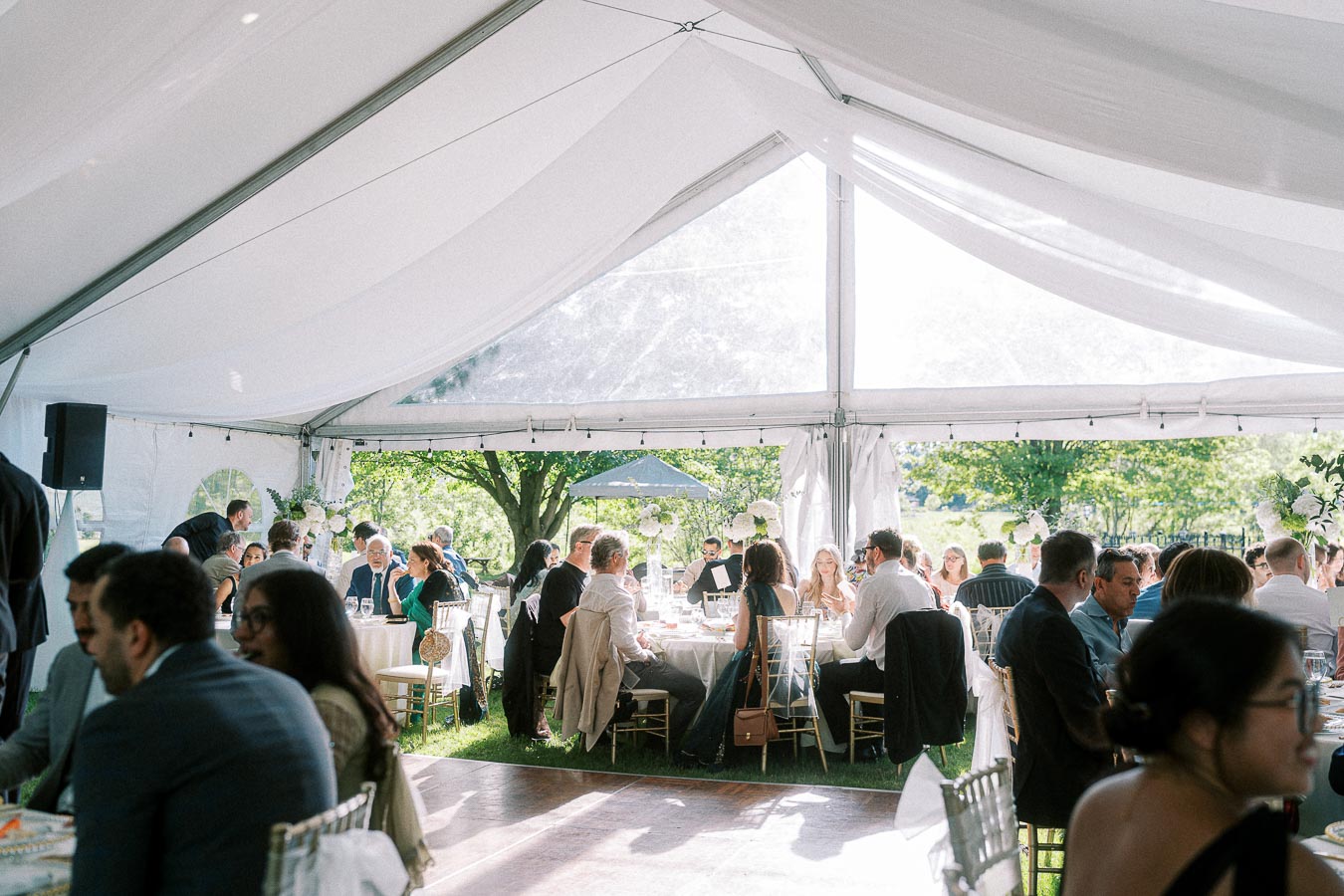 Outdoor wedding reception under a white tent with guests seated at round tables, surrounded by lush greenery and soft natural light.
