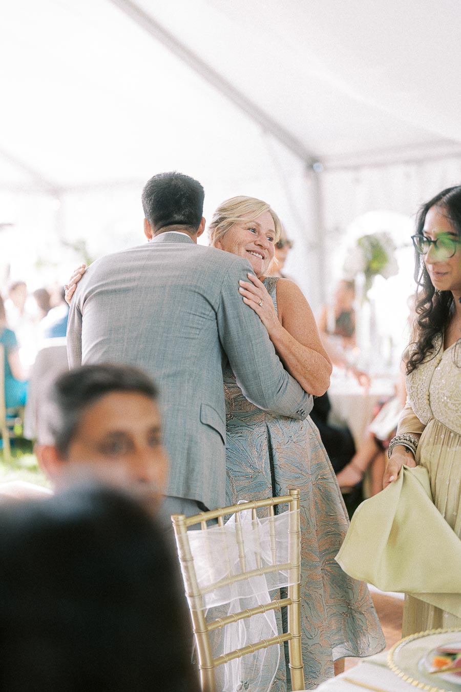 A warm hug between guests at a lively outdoor wedding reception under a white tent, featuring elegantly dressed individuals enjoying the celebration.