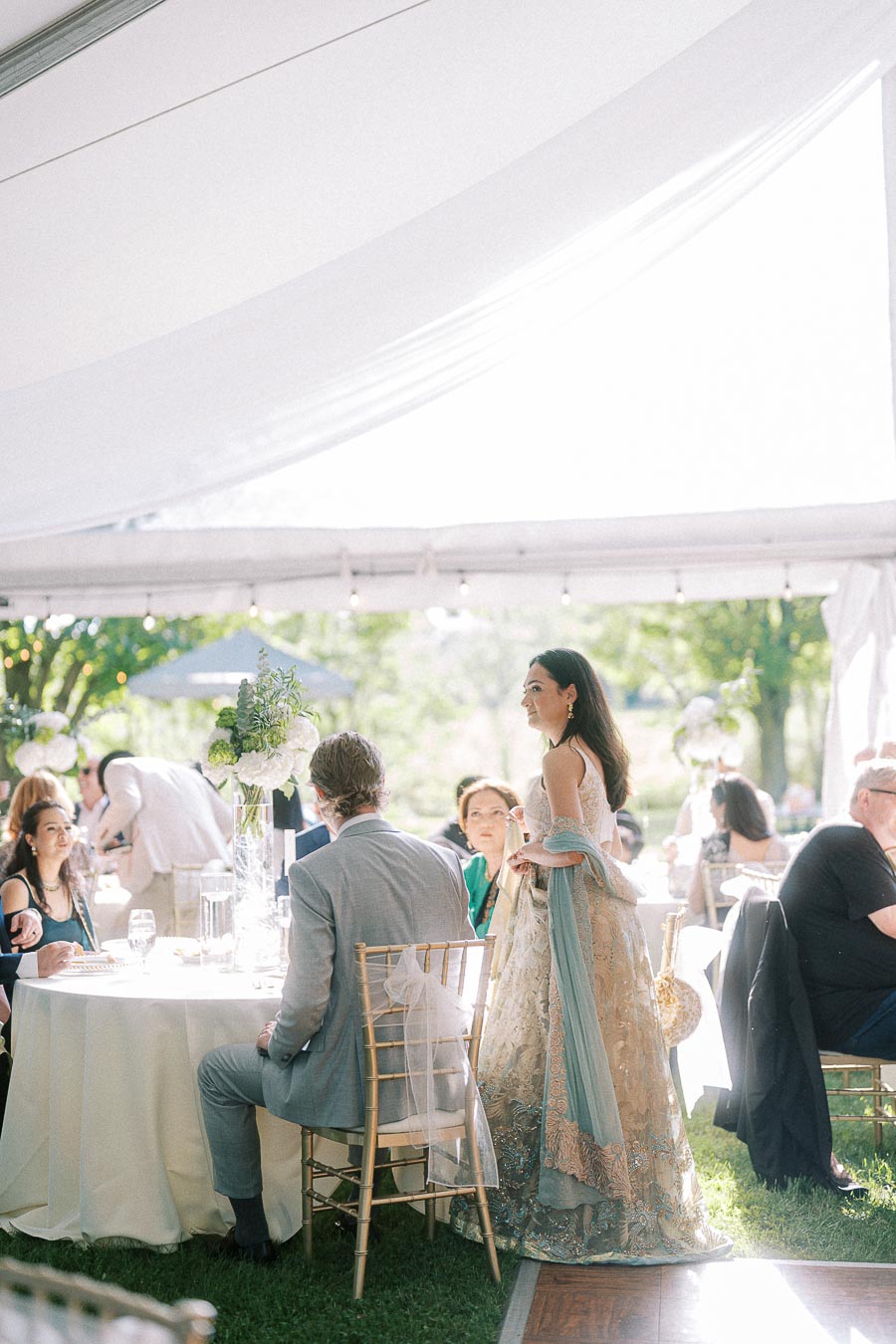 Elegant garden wedding reception under a white tent with guests seated at round tables; a woman in a decorative gown standing and socializing.