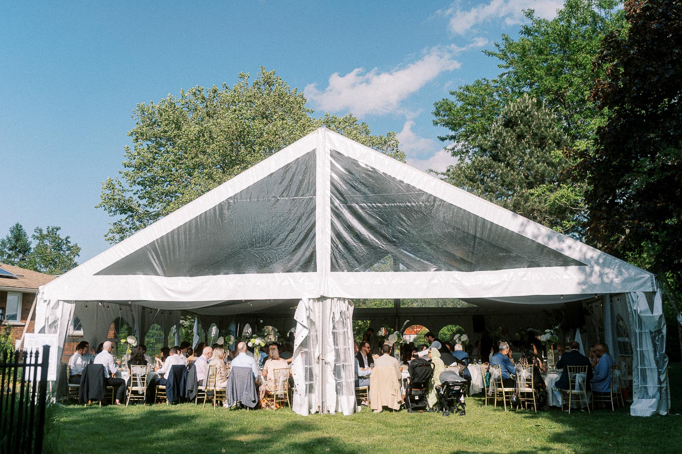 Outdoor wedding reception in a large white tent with guests seated at tables surrounded by lush greenery and a clear blue sky.