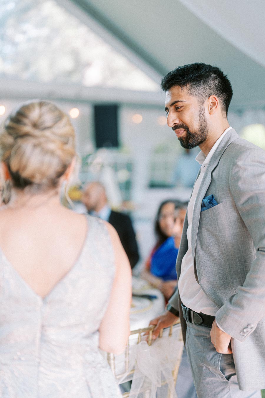 Man in a gray suit smiling and conversing with a woman at a formal event, with elegantly dressed guests seated in the blurred background.
