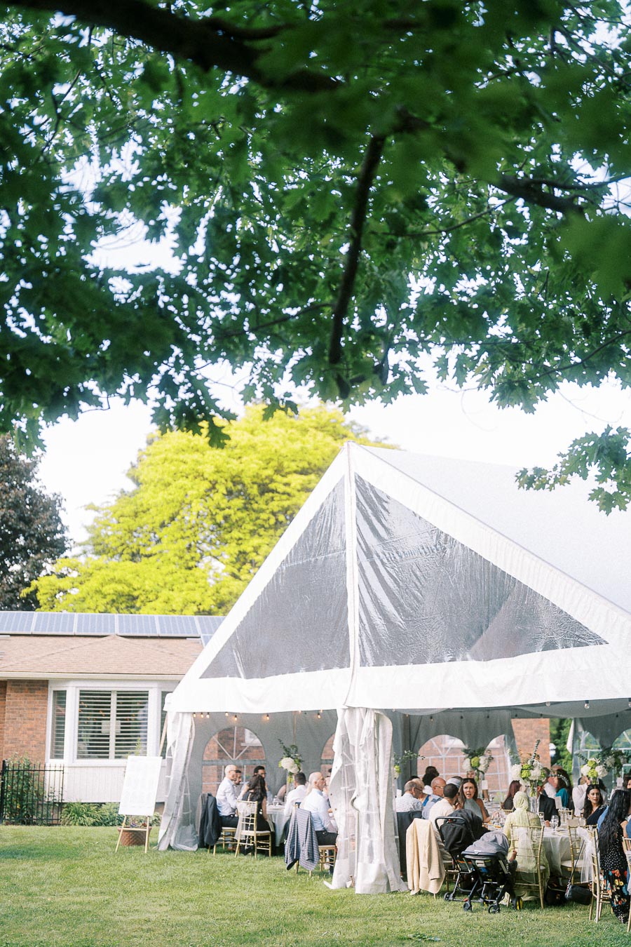 Outdoor wedding reception under a clear marquee tent surrounded by lush greenery and guests seated at elegantly decorated tables, set against a backdrop of a brick building with solar panels.