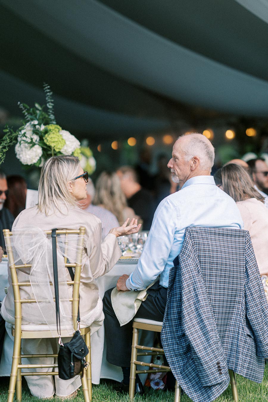 Elderly couple engaged in conversation at a garden wedding reception, seated on gold chairs under a tent with elegant decor and floral arrangements.