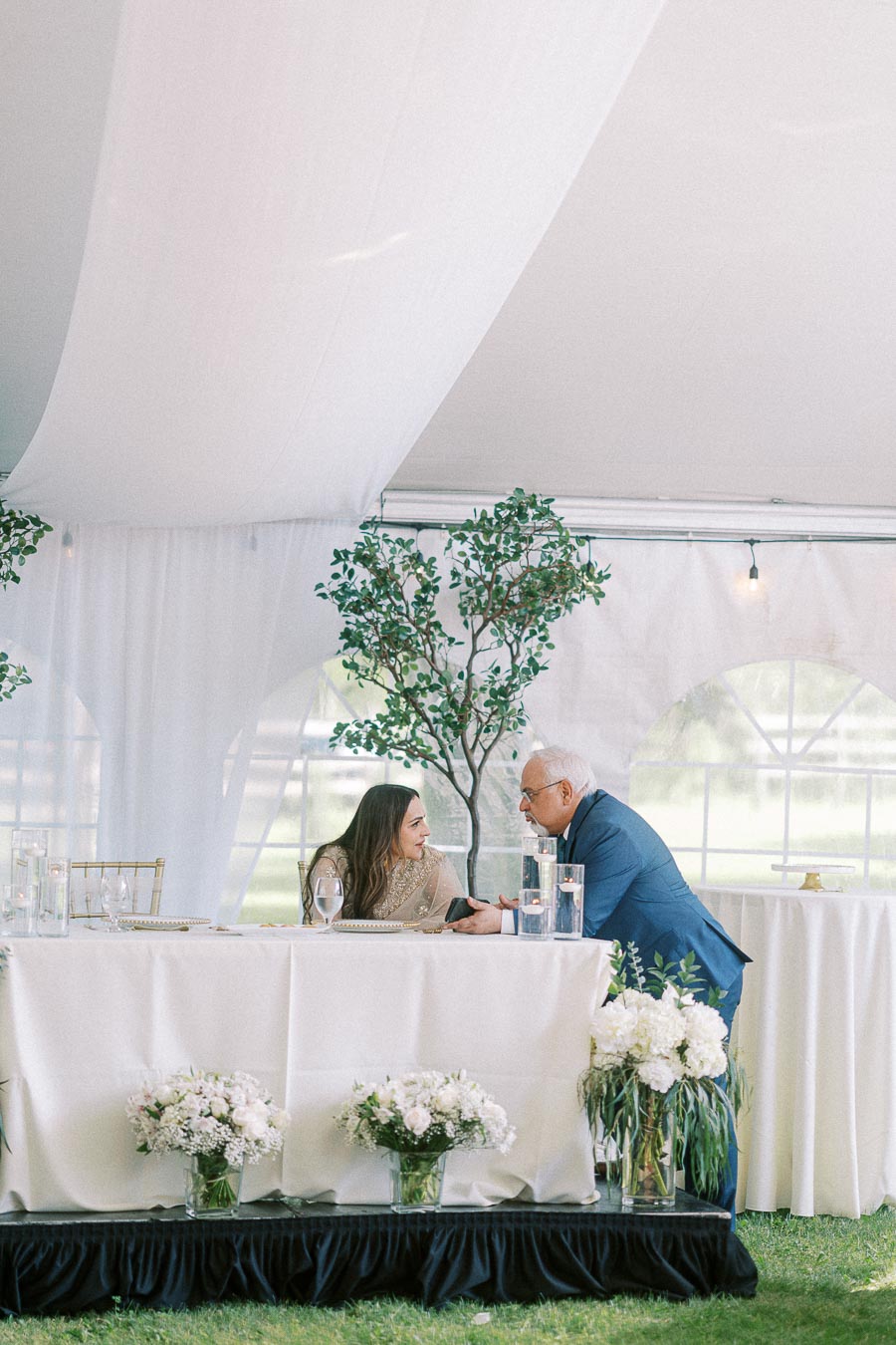 Elegant wedding reception under a tent with a woman in traditional attire and a man in a blue suit conversing at a beautifully decorated table with floral arrangements and candles.