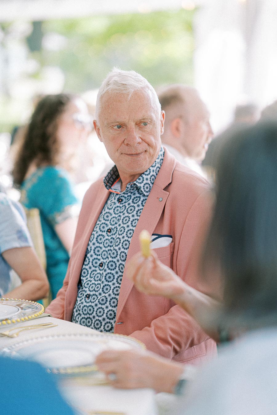 Elderly man in a pink jacket with a patterned shirt, seated at a formal dining event, engaging in conversation with other guests.