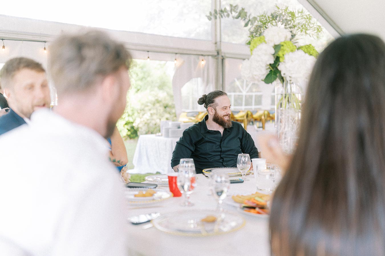 A cheerful group of people sitting around a decorated table at an outdoor event or wedding, with a bearded man in a black shirt smiling. Elegant table setting with glassware and floral centerpiece visible.
